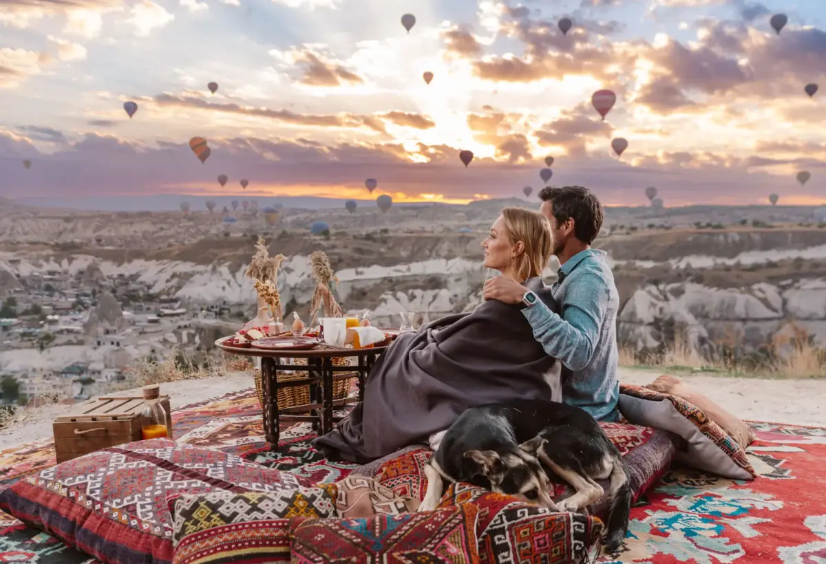 Two travellers at a sunrise breakfast laid out on kilim rugs above a Cappadocian valley, balloons in the sky behind them.